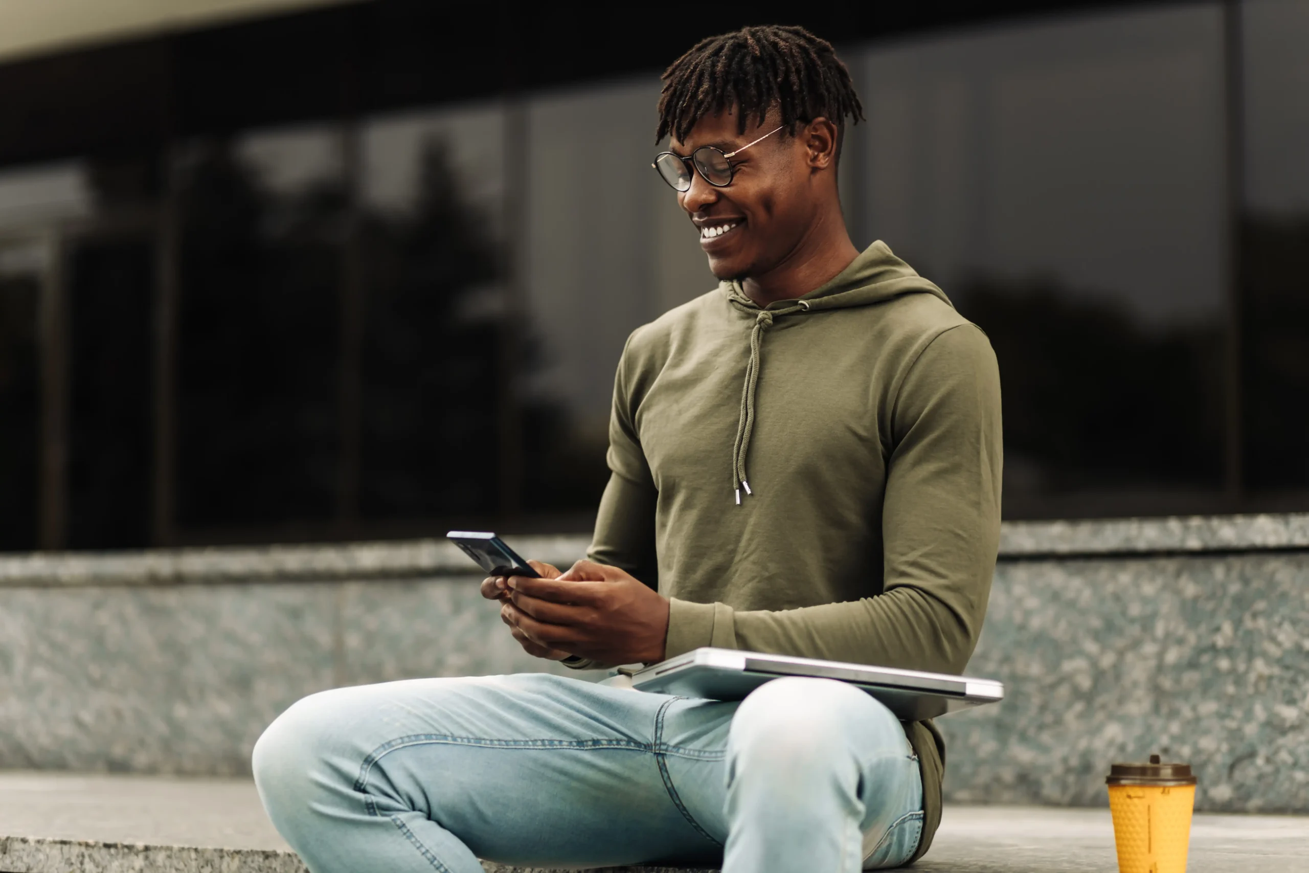 Young man sitting outside a building with coffee smiling while looking at his cell phone