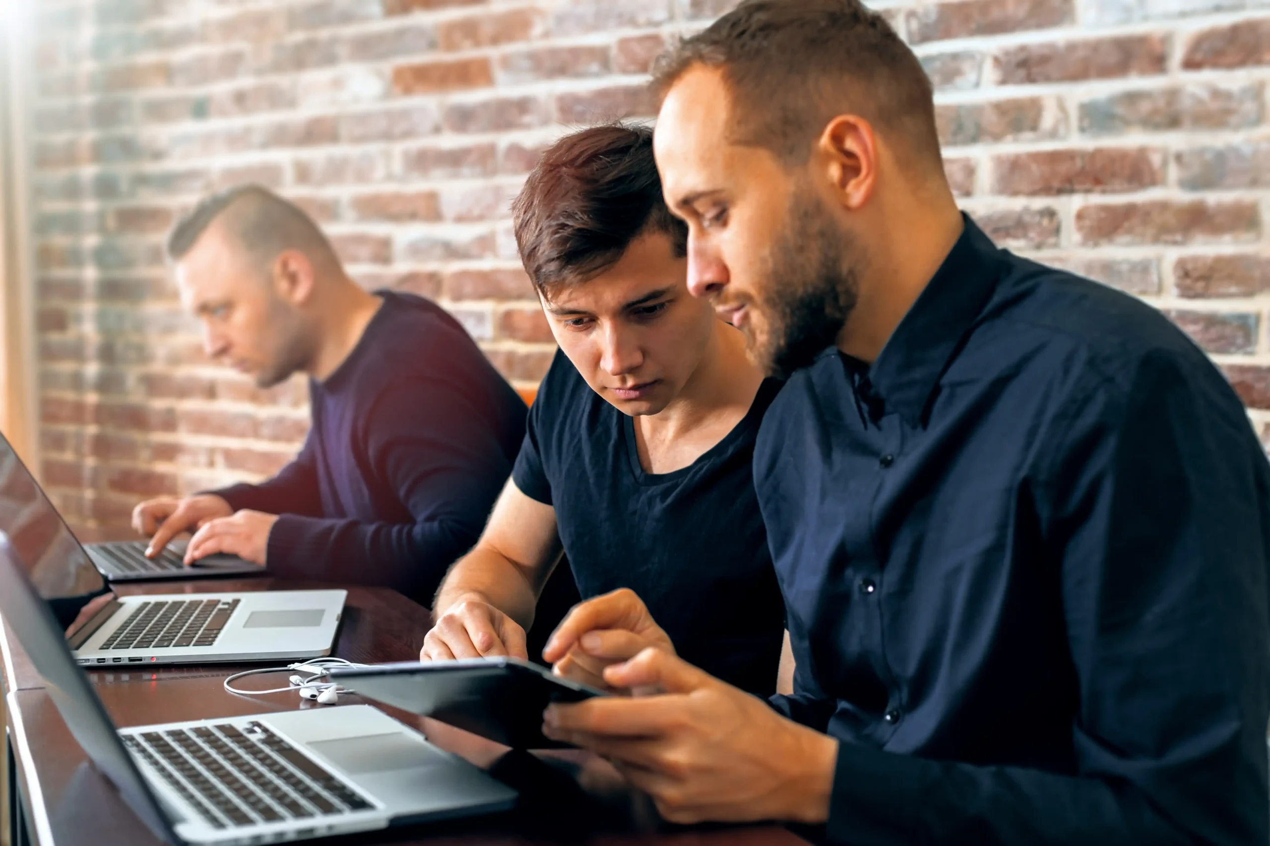 Three men at shared desk working together on devices to solve a problem