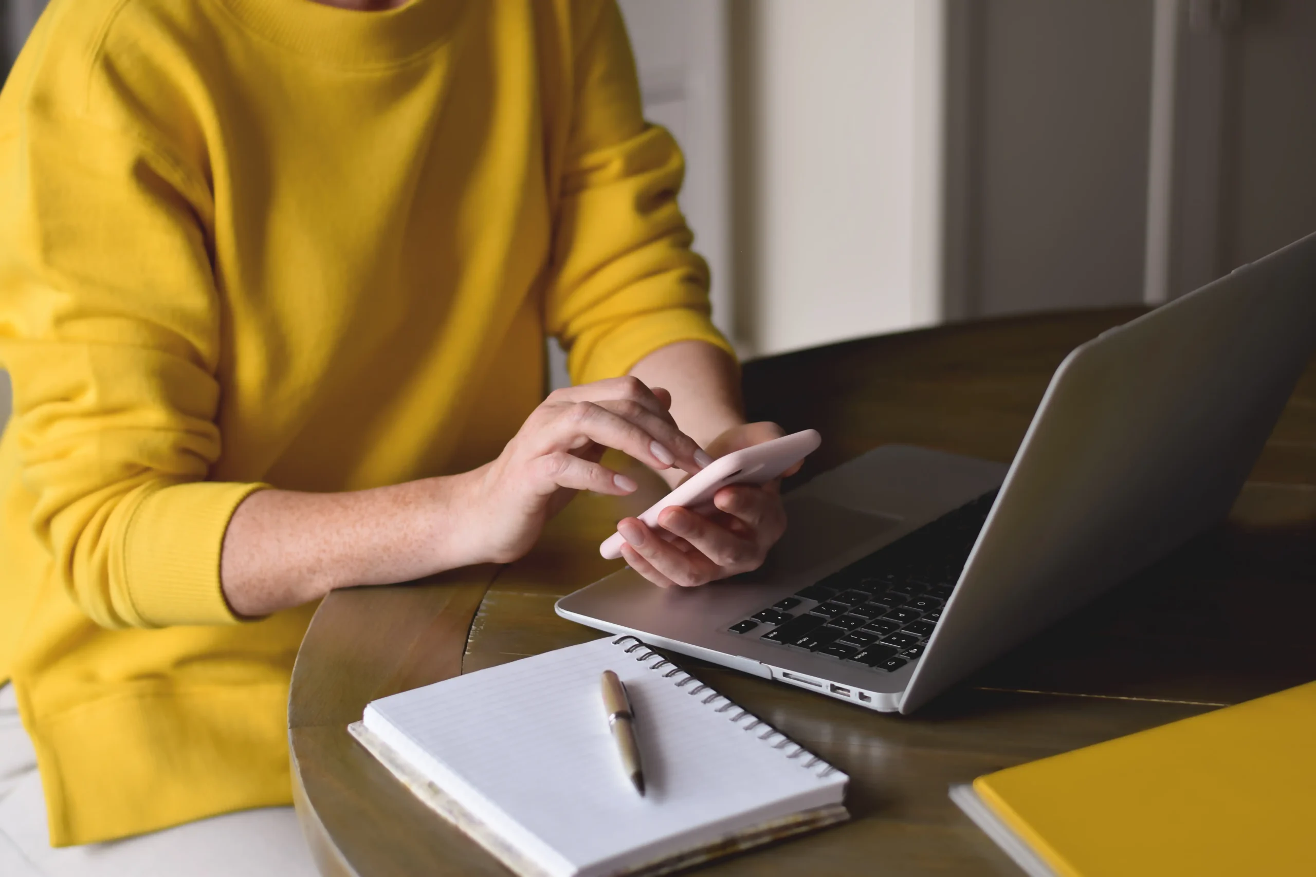 Woman in yellow sweater at desk on her phone and laptop